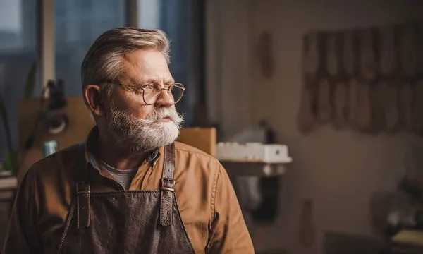 A white-bearded craftsman in a leather apron looking out the window of his workshop, round glasses on, rows of leather hanging on the wall behind him.
