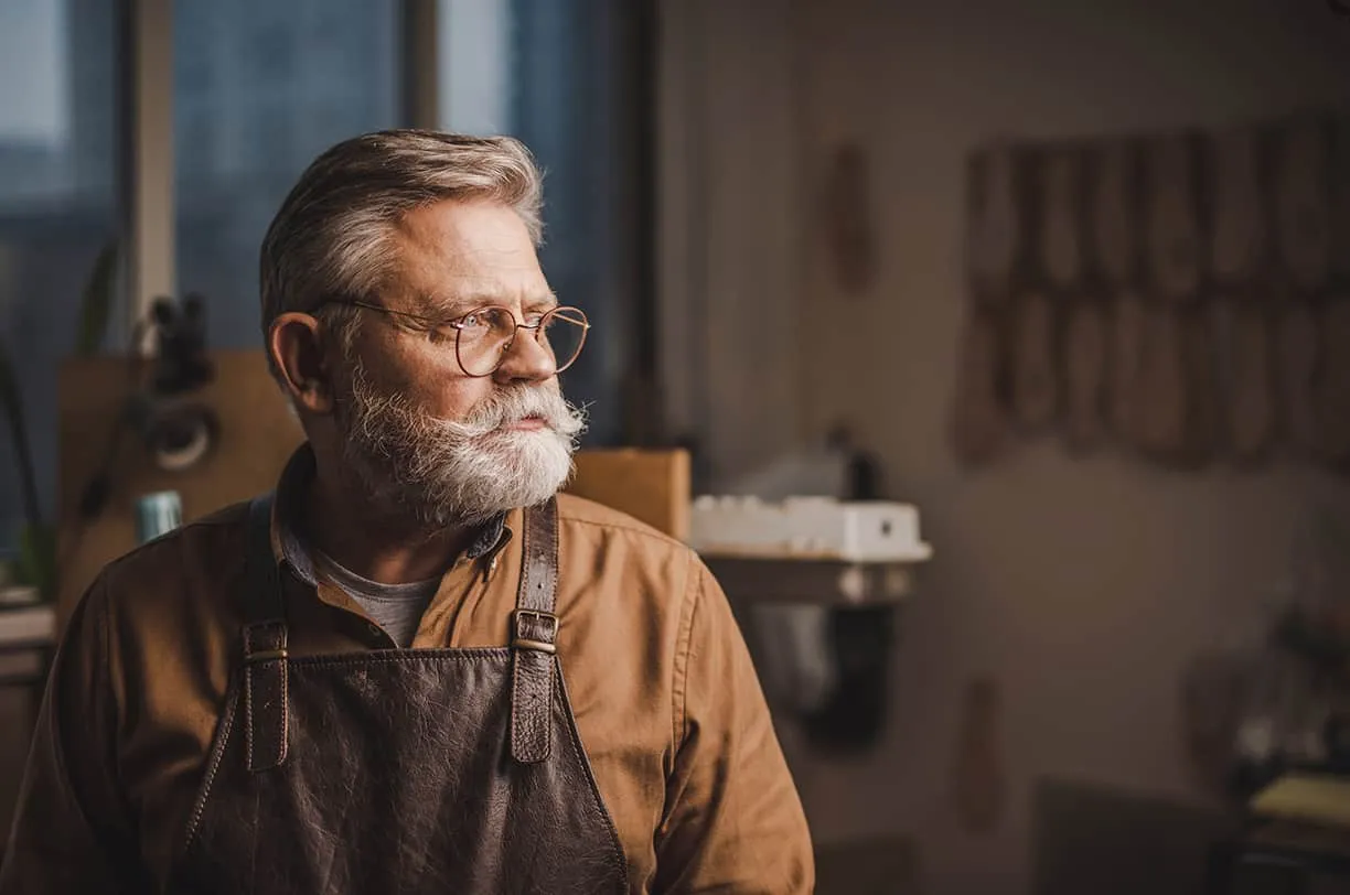 A white-bearded craftsman in a leather apron looking out the window of his workshop, round glasses on, rows of leather hanging on the wall behind him.